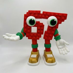 May include: A whimsical red number 67 figure with large googly eyes, white hands, and gold shoes. The figure has green and red accents on its arms and legs, and is posed with its hands open. The figure is a fun, playful design.