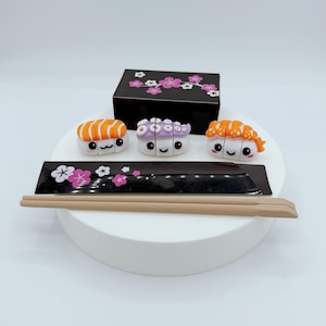 Op de afbeelding: Een sushi-thema set met een zwarte doos en een zwart dienblad, beide versierd met roze bloemmotieven. Drie sushi-stukken met schattige gezichten worden getoond, samen met een paar lichtbruine eetstokjes. De sushi-stukken hebben oranje, paarse en witte elementen.