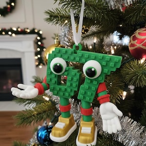 May include: A whimsical Christmas tree ornament shaped like the number 67, constructed from green blocks. It features large googly eyes, red and green arms and legs, and gold shoes. The ornament hangs from a white ribbon on a decorated Christmas tree.