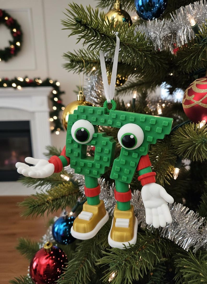 May include: A whimsical Christmas tree ornament shaped like the number 67, constructed from green blocks. It features large googly eyes, red and green arms and legs, and gold shoes. The ornament hangs from a white ribbon on a decorated Christmas tree.