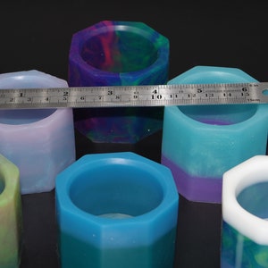 Op de afbeelding: Zes zeshoekige kaarshouders van hars in verschillende kleuren, waaronder blauw, groen, paars en wit. De houders zijn ongeveer 5 cm in diameter en 5 cm hoog.