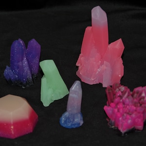 May include: A collection of colourful crystal formations in shades of purple, pink, green, and blue. The crystals vary in size and shape, with some resembling clusters and others single points. The crystals are displayed against a black background.