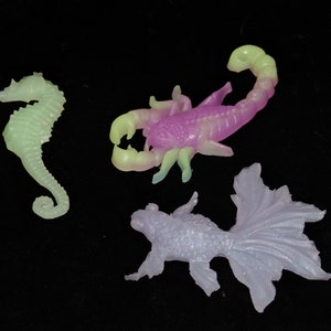 May include: Three glow-in-the-dark plastic sea creatures: a purple scorpion, a green seahorse, and a white fish.