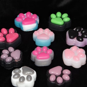 May include: Assortment of colourful paw print shaped soaps. The soaps are in various colours, including pink, green, blue, purple, and grey. Some of the soaps have glitter on them.