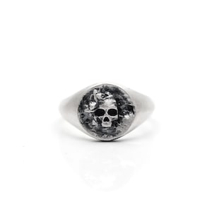 May include: A silver signet ring with a skull design. The ring has a rounded face with a detailed skull carving. The metal has a slightly textured appearance. The ring is set against a white background.