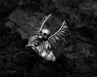 May include: A silver ring with a skull and bat wings design. The ring is made of metal and has a detailed design.