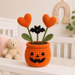 May include: A crocheted orange jack-o'-lantern pot containing two orange heart-shaped flowers and a small black bat on green stems. The pot features a black stitched jack-o'-lantern face with round eyes, a triangular nose, and a wide, jagged grin.