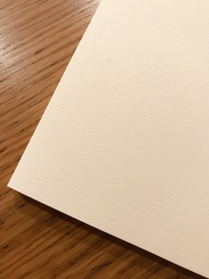 May include: A corner of a cream-colored, textured paper sheet rests on a wooden surface. The paper has a subtle, horizontal ribbed pattern. The wood grain is visible in the background, creating a contrast in texture and color.