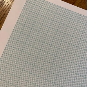 May include: A sheet of graph paper with a light blue grid pattern on a white background. The grid is composed of evenly spaced horizontal and vertical lines, creating a series of small squares. The paper is slightly angled, showing a corner.