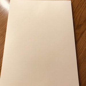 May include: A rectangular sheet of off-white paper, likely for writing or drawing, is centered on a wooden surface. The paper has a smooth, matte finish and a slight texture. The wood grain is visible.