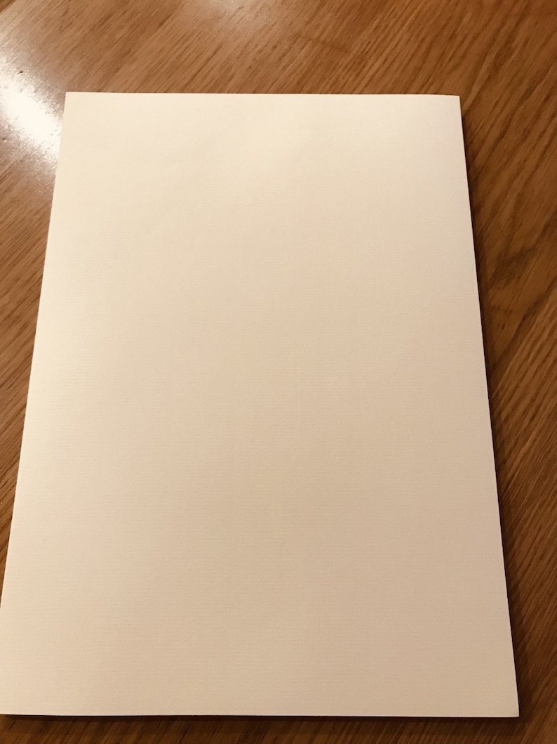 May include: A rectangular sheet of off-white paper, likely for writing or drawing, is centered on a wooden surface. The paper has a smooth, matte finish and a slight texture. The wood grain is visible.