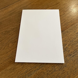 May include: A plain white rectangular card, possibly for a message or note, lies on a wooden surface. The card is centrally positioned, occupying much of the image, with the wood grain visible around it.