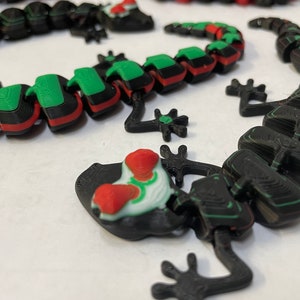 May include: Three 3D printed lizards, one black with green and red accents, one black with green accents, and one red with green accents.