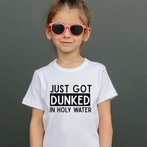 May include: A white t-shirt with the text "JUST GOT DUNKED IN HOLY WATER" in black. The shirt is worn by a person wearing pink sunglasses. The background is a solid grey colour.