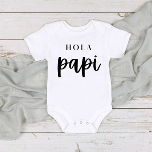 May include: White baby onesie with the words "HOLA papi" printed in black. The onesie has short sleeves and snaps at the bottom. The text is in a bold, modern font. The onesie is laying on a light gray fabric and a white wooden surface.