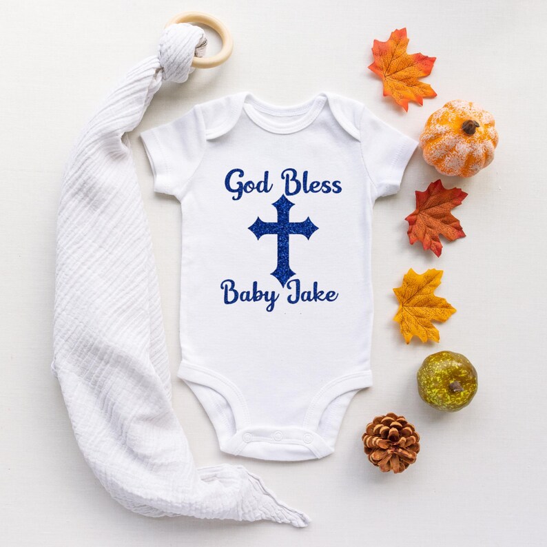 Op de afbeelding: Witte babybody met de blauwe tekst "God Bless Baby Jake". Een blauw kruis is gecentreerd onder de woorden. De body is omgeven door herfstdecoraties, waaronder bladeren, een pompoen en een dennenappel.