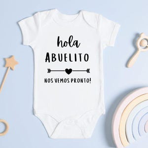 May include: White baby onesie with the text "hola ABUELITO nos vemos pronto!" in black. The onesie is surrounded by wooden toys, including star-shaped wands, a rattle, and a rainbow arch, all on a light blue surface.
