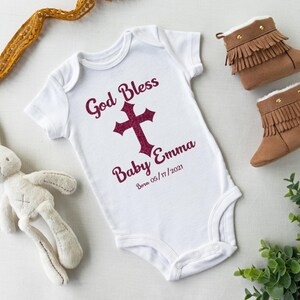 Op de afbeelding: Witte babybody met de tekst "God Bless Baby Emma Born 05/17/2021" en een bordeauxrode kruisafbeelding. De body ligt naast een paar bruine babyboots met franjes en een knuffelkonijn.