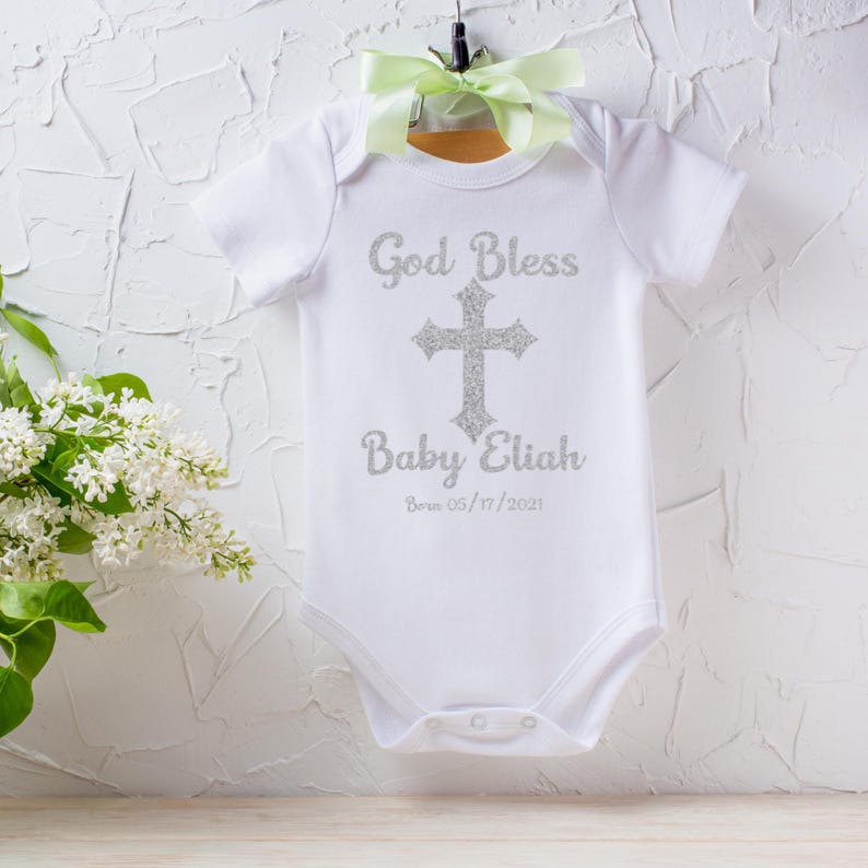 Op de afbeelding: Witte babybody met korte mouwen, met de tekst "God Bless Baby Eliah" en een zilveren glitterkruis. De datum "Born 05/17/2021" is eronder gedrukt. De body hangt aan een houten hanger met een groen lint.