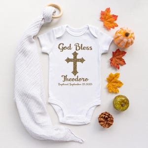 Op de afbeelding: Witte babybody met gouden glittertekst "God Bless" boven een kruis en de naam "Theodoro" met de doopdatum. De afbeelding bevat herfstbladeren en een houten bijtring.