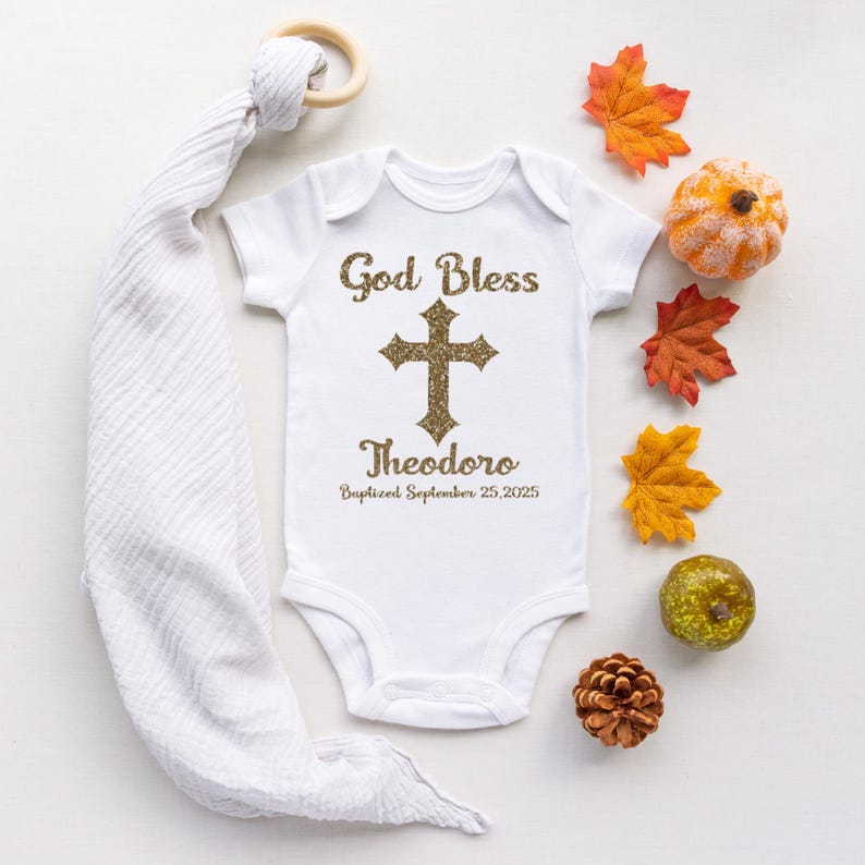 Op de afbeelding: Witte babybody met gouden glittertekst "God Bless" boven een kruis en de naam "Theodoro" met de doopdatum. De afbeelding bevat herfstbladeren en een houten bijtring.