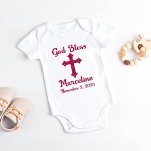 Op de afbeelding: Witte babybody met de tekst "God Bless" boven een glitterroze kruis en de naam "Marceline" met de datum "3 november 2024". De afbeelding bevat ook babyschoenen en een houten speelgoed.