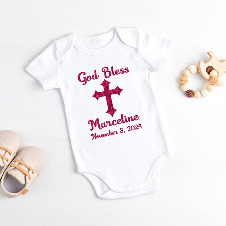 Op de afbeelding: Witte babybody met de tekst "God Bless" boven een glitterroze kruis en de naam "Marceline" met de datum "3 november 2024". De afbeelding bevat ook babyschoenen en een houten speelgoed.