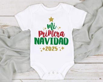 May include: White baby onesie with the text "Mi Primera Navidad 2025" in red, green, and gold. The design includes a gold star and sparkles. The onesie is laid on a light gray fabric and a white wooden surface.