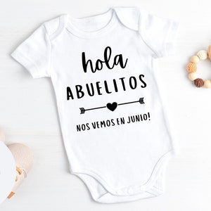 May include: White baby onesie with black text that reads "hola abuelitos nos vemos en junio!" with a heart and arrow design. A wooden baby toy and a logo are also visible.