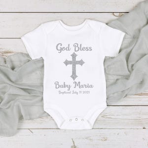 Op de afbeelding: Witte babybody met de tekst "God Bless" boven een zilveren kruis en "Baby Maria" eronder. De datum "Baptized July 11 2021" staat onder de naam gedrukt. De body ligt op een lichtgrijze stof.
