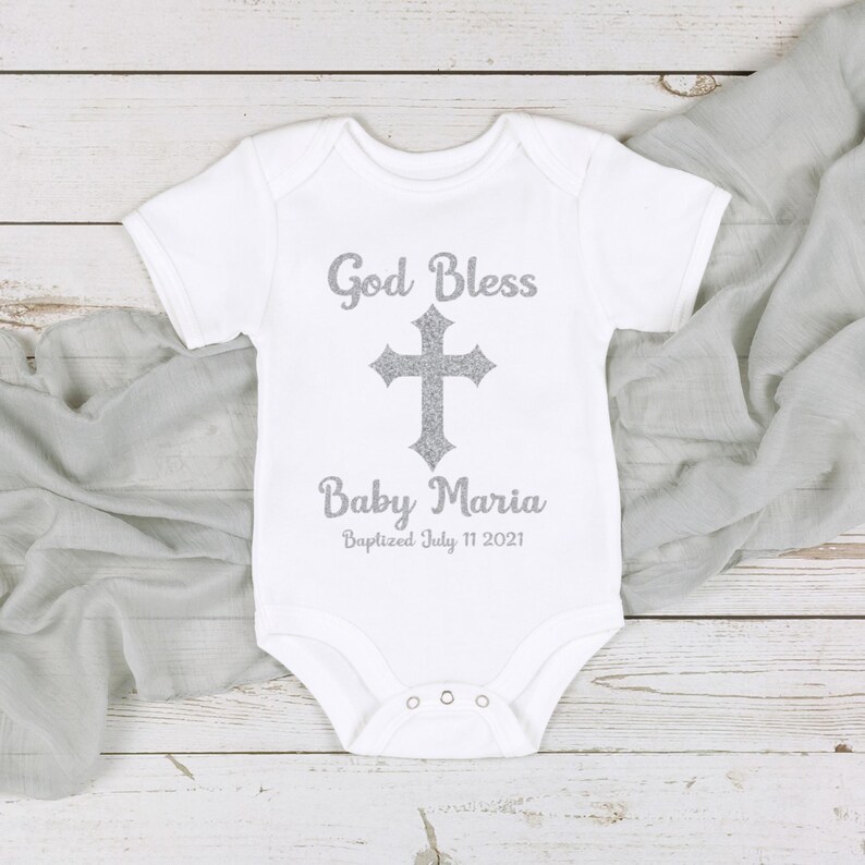 Op de afbeelding: Witte babybody met de tekst "God Bless" boven een zilveren kruis en "Baby Maria" eronder. De datum "Baptized July 11 2021" staat onder de naam gedrukt. De body ligt op een lichtgrijze stof.