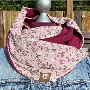 Puede incluir: Una bufanda infinita de doble cara rosa y burdeos con estampado floral y una etiqueta de cuero en forma de corazón.