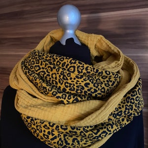 May include: Mustard yellow infinity scarf with a leopard print pattern. The scarf is displayed on a mannequin and features a double-layered design. The leopard print is black on a tan background. The scarf is made of a soft, textured fabric.