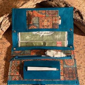 Könnte beinhalten: Tealfarbene Leder-Tabaktasche zum Selberrollen mit Reißverschlussfach und gemustertem Stofffutter. Die Tasche hat ein transparentes Plastikfenster und einen Trageriemen.