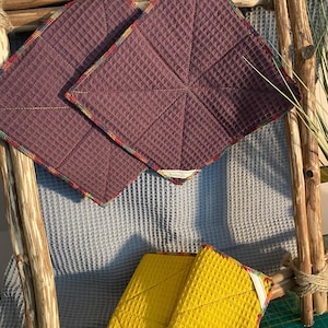 Op de afbeelding: Twee vierkante, pruimkleurige wafeldoeken met kleurrijke rand worden getoond. Daaronder is een stapel gele doeken met dezelfde wafelstof en rand zichtbaar. De doeken zijn gerangschikt op een rustiek houten frame.