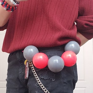 Op de afbeelding: Een persoon draagt een bordeauxrode trui en een zwarte jeans met een ketting die aan hun riem is bevestigd. De ketting heeft een sleutel eraan. De persoon draagt een ketting en verschillende kralenarmbanden. De persoon draagt een riem met een rij rode en grijze ballen die eraan zijn bevestigd.