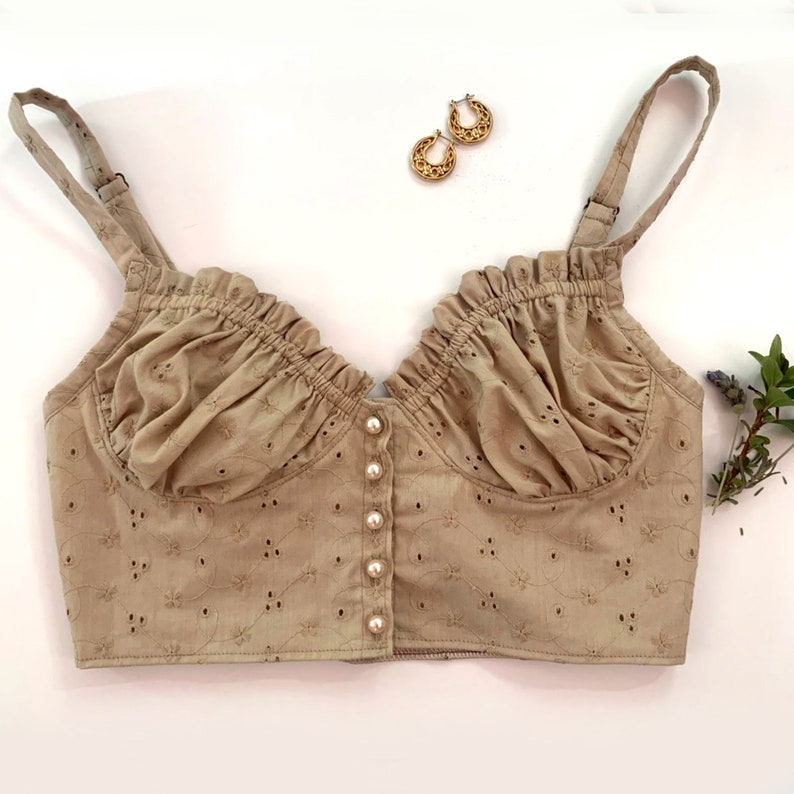 May include: A beige, cropped, sleeveless top with a ruffled neckline and a button-up front. The top is made of a delicate, eyelet fabric.