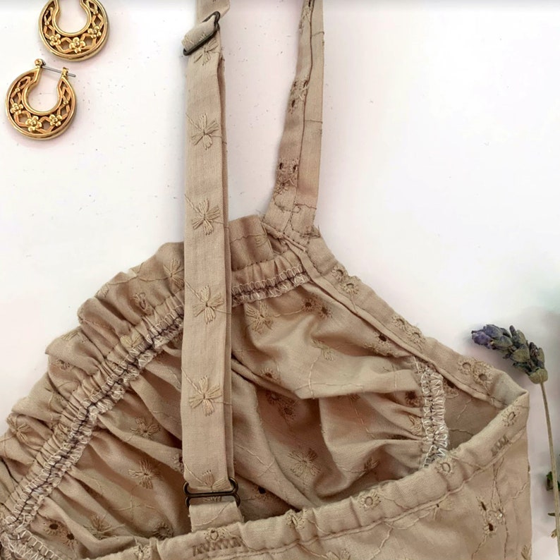 May include: A beige sleeveless top with embroidered floral details and adjustable straps. The top has a gathered neckline and a relaxed fit.