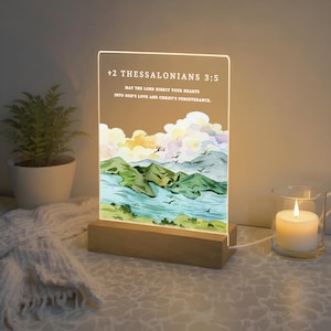 May include: An illuminated acrylic panel with a wooden base, displaying a watercolor landscape and the text "+2 THESSALONIANS 3:5" with a Bible verse. The image includes a lit candle in a glass and a small potted plant. The light is powered by a cable.