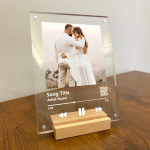 May include: A clear acrylic photo frame with a wooden stand. The frame features a photo of a couple and a music player design with the text "Song Title" and "Artist Name".