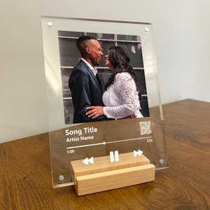 May include: A clear acrylic photo frame with a wood stand. The frame features a photo of a couple and a music player design with the text "Song Title" and "Artist Name".