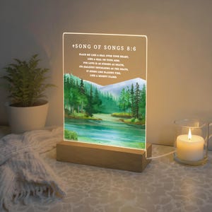 May include: An illuminated acrylic panel featuring the text "+SONG OF SONGS 8:6" and a watercolour landscape of a lake and trees. The panel is set on a wooden base. A lit candle in a glass holder is placed nearby.