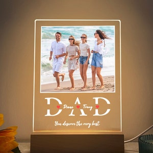 May include: A white illuminated acrylic sign with the word "DAD" in large white letters. The sign features a photo of a family of four on a beach. The text "You deserve the very best." is below the photo. The names "Diane" and "Tracy" are written in smaller letters above the hearts.