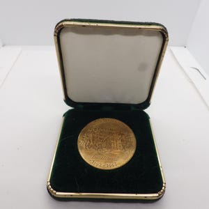 May include: A circular gold-colored commemorative coin featuring an embossed scene and the text United States Mint Bicentennial 1792-1992, displayed inside a square presentation case with a dark green velvet interior and a white satin lid lining.