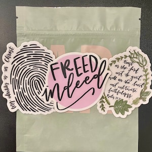 Puede incluir: Tres pegatinas sobre un fondo verde claro. La primera pegatina es una huella dactilar con el texto "my identity is in Christ." La segunda pegatina es rosa con el texto "FREED indeed." La tercera pegatina es una corona de hojas verdes con el texto "trust in the Lord and to do good. Dwell in the land and cultivate faithfulness. Psalm 37:3."