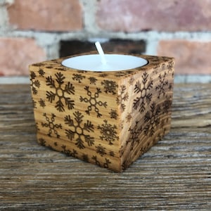 Può includere: Un portacandele in legno con un motivo a fiocco di neve. Il portacandele è quadrato e ha una piccola apertura al centro per una candela da tè.