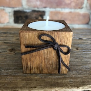 Può includere: Un piccolo portacandele in legno con un fiocco in pelle marrone. Il portacandele è realizzato in legno naturale e ha una forma quadrata. È perfetto per contenere una candela da tè.