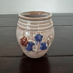 Può includere: Un piccolo vaso in ceramica con una base marrone chiaro e strisce orizzontali bianche vicino alla parte superiore. Il vaso presenta disegni floreali blu e marroni. Il vaso è appoggiato su una superficie marrone scuro.