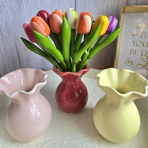 Può includere: Un bouquet di tulipani di legno colorati in un vaso rosso, con foglie verdi. Altri due vasi in ceramica, uno rosa e uno giallo, sono in primo piano. I tulipani sono di vari colori, tra cui arancione, rosso e giallo.