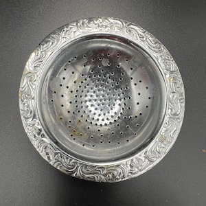 Pode incluir: Um coador de chá prateado com um padrão floral decorativo e ornamentado ao redor da borda. O coador tem uma tigela perfurada para filtrar as folhas de chá. O coador de chá é feito de metal e tem formato circular.